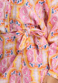 Multikleurige stof met ingewikkelde bloem- en paisleypatronen in roze, oranje en gele tinten; heeft een knopen aan de voorkant en een strikceintuur.