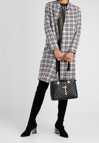 Nainen, jolla on päällään ruudullinen takki ja mustat polvipituiset saappaat, pitää kädessään mustaa tikattua laukku, jossa on kultaketjuolkaimet ja tupsu.