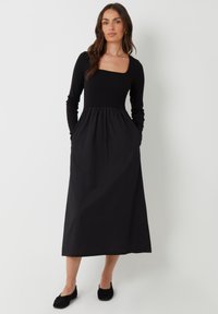Vestido negro de manga larga con escote cuadrado, corsé ajustado y falda acampanada. Presenta bolsillos laterales y una textura suave.