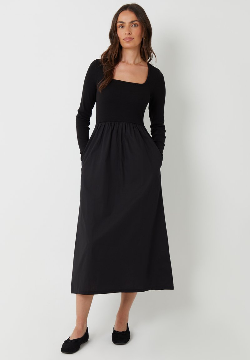 Vestido negro de manga larga con escote cuadrado, corsé ajustado y falda acampanada. Presenta bolsillos laterales y una textura suave.