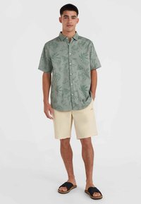 Kurzärmliges, hellgrünes Hemd mit Knopfleiste, verziert mit Blatt- und geometrischen Mustern, kombiniert mit beigen Shorts und schwarzen Sandalen.