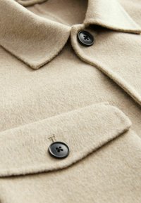 Col de manteau en laine beige et poche à rabat avec deux boutons noirs visibles à quatre trous, montrant la texture et les détails de couture.