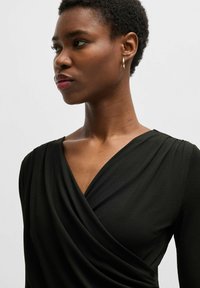 Black wrap dress with a plunging neckline, draped texture, and long sleeves. Model features short hair and gold hoop earrings.