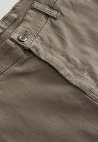 Beige kokvilnas chinos ar strukturētu audumu, ar pogu aizdari un jostas cilpu. Tuvplāns skats uz priekšējo paneli.