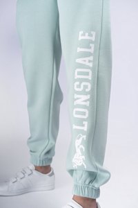 Mätovo zelené tepláky s elastickými manžetami, so bielym brandingom "LONSDALE" a grafikou leva na boku. V kombinácii s bielymi teniskami.