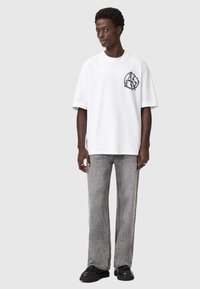 Valkoinen suurikokoinen t-paita, jossa on musta graafinen logo vasemmassa rinnassa, yhdistettynä haalistuneisiin harmaisiin leveälahkeisiin farkkuihin ja mustiin kenkiin.