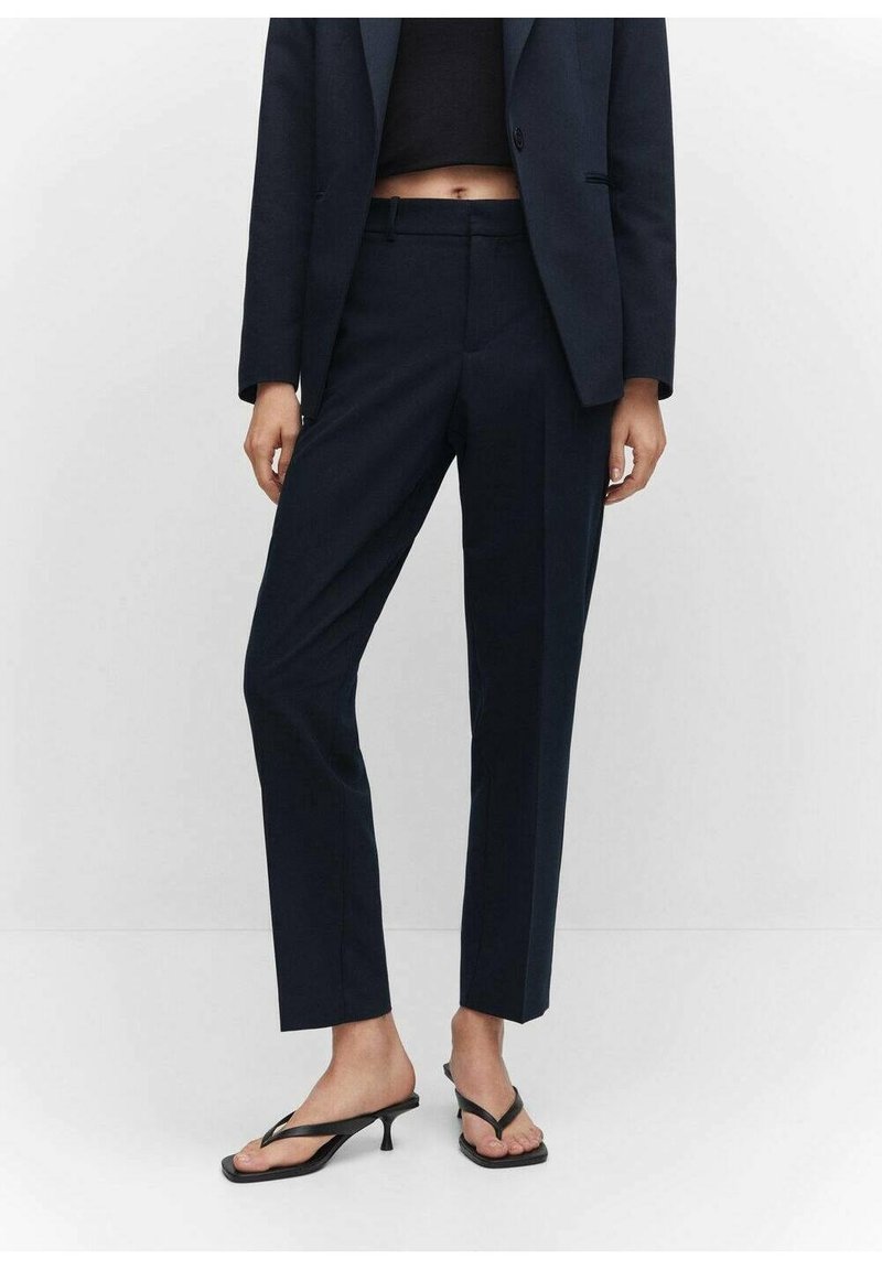 Femme portant un pantalon bleu marine sur mesure, un blazer assorti, un top noir court et des sandales à talons noires, debout devant un fond blanc.