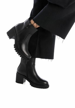 Botas negras hasta el tobillo con un tacón grueso, parte superior de cuero suave, cierre de cremallera lateral y suela de goma con un patrón estriado.