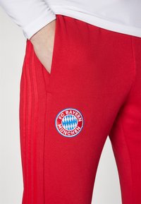 Røde treningbukser med en tettsittende passform, utstyrt med et brodert FC Bayern München-logo. Inkluderer sidelommer og tre vertikale striper.
