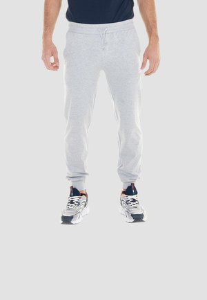 Man draagt lichtgrijze joggerbroeken, donkerblauw shirt en witte hardloopschoenen met zwarte en oranje accenten, staand op een grijze achtergrond.