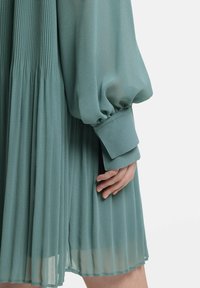 A pleated dress in soft teal fabric, featuring voluminous long sleeves with cuffs, showcasing a flowing design and delicate texture.