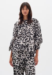Femeie îmbrăcată cu un set potrivit de bluză și pantaloni negri și albi cu imprimeu abstract, cu mâneci puff până la trei sferturi, stând în fața unui fundal alb simplu.