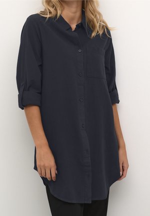 Femme portant une chemise boutonnée bleu marine foncé à manches longues, avec les manches retroussées et une poche poitrine, debout devant un fond clair uni.
