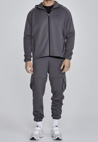 Grå zip-up jacka med huva, i kombination med matchande cargobyxor. Båda tillverkade av mjukt, elastiskt material. Har sidofickor och elastiska muddar.