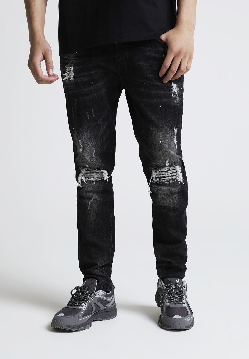 Personne portant un jean noir déchiré et des baskets grises, debout devant un fond blanc uni.