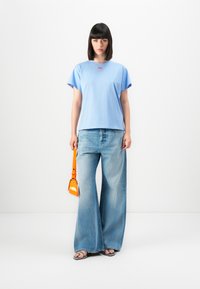 Jeune femme aux cheveux noirs raides, portant un T-shirt bleu clair et un jean à jambes larges, tenant un petit sac à main orange, se tenant sur fond blanc.