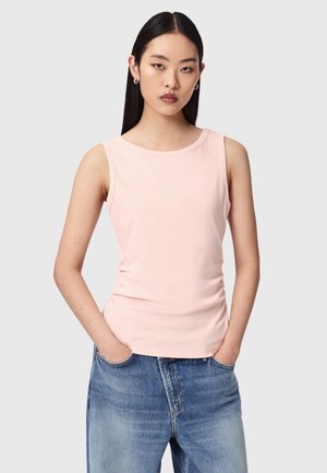 Femme aux cheveux noirs raides portant un débardeur rose pâle et un jean bleu, debout les mains dans les poches devant un fond uni.