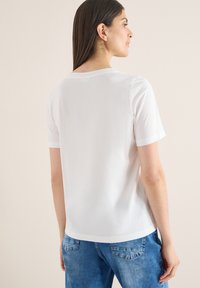 Femme de dos portant un t-shirt blanc uni à manches courtes et un jean bleu sur un fond beige.