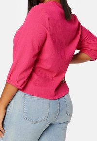 Blouse rose texturée avec manches bouffantes trois-quarts, associée à un jean en denim bleu clair avec une poche arrière.