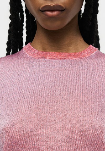 Κοντινό πλάνο ενός ατόμου με σκούρα πλεγμένα μαλλιά που φοράει ροζ πλεκτό πουλόβερ με στρογγυλή λαιμόκοψη.