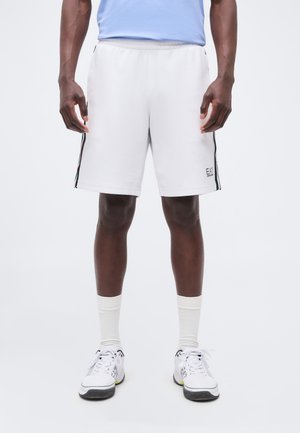 Mees kannab valgeid spordipükse mustade, roheliste ja punaste küljeribadega, valgeid sokke ja valgeid tossusid, seisab heleda tausta ees.