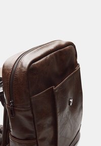 Zaino in pelle marrone con una texture liscia, chiusura superiore con zip e tasca frontale con un accento argentato.
