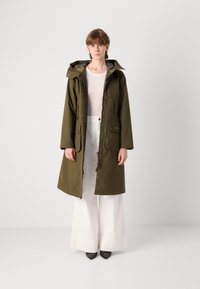 Olijfgroene waterdichte parka met een capuchon, knopen aan de voorkant, twee grote zakken en een kleurrijke binnenvoering, gedragen over witte wijde pantalons.