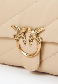 Μπεζ κουβάρια τσάντα με χρυσό μεταλλικό κούμπωμα που απεικονίζει δύο πουλιά που κοιτάζονται, με ανάγλυφο το "PINKO" και κυκλικό διακοσμητικό.