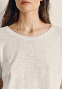 Žena s dlhými hnedými vlasmi nosiaca biele tričko s plochým výstrihom, viditeľná od ramien po ústa na jednoduchom svetlom pozadí.