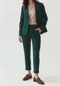Completo verde in lana con blazer su misura e pantaloni a sigaretta, abbinato a un top beige. Mocassini in pelle marrone completano l'outfit.