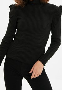 Czarna, prążkowana bluzka z długim rękawem, z wysokim kołnierzem, falbankami na ramionach i ozdobnymi guzikami w złotym odcieniu na mankietach. Miękka tekstura.