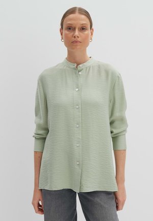 Helles grünes Hemd mit Stehkragen, langen Ärmeln und strukturiertem Stoff. Weiße Knöpfe und eine entspannte Passform. Kombiniert mit grauen Hosen.