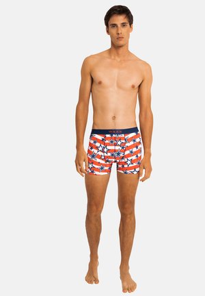 Jeune homme debout pieds nus portant un boxer rouge, blanc et bleu à motifs d’étoiles avec une ceinture bleu marine sur un fond uni.