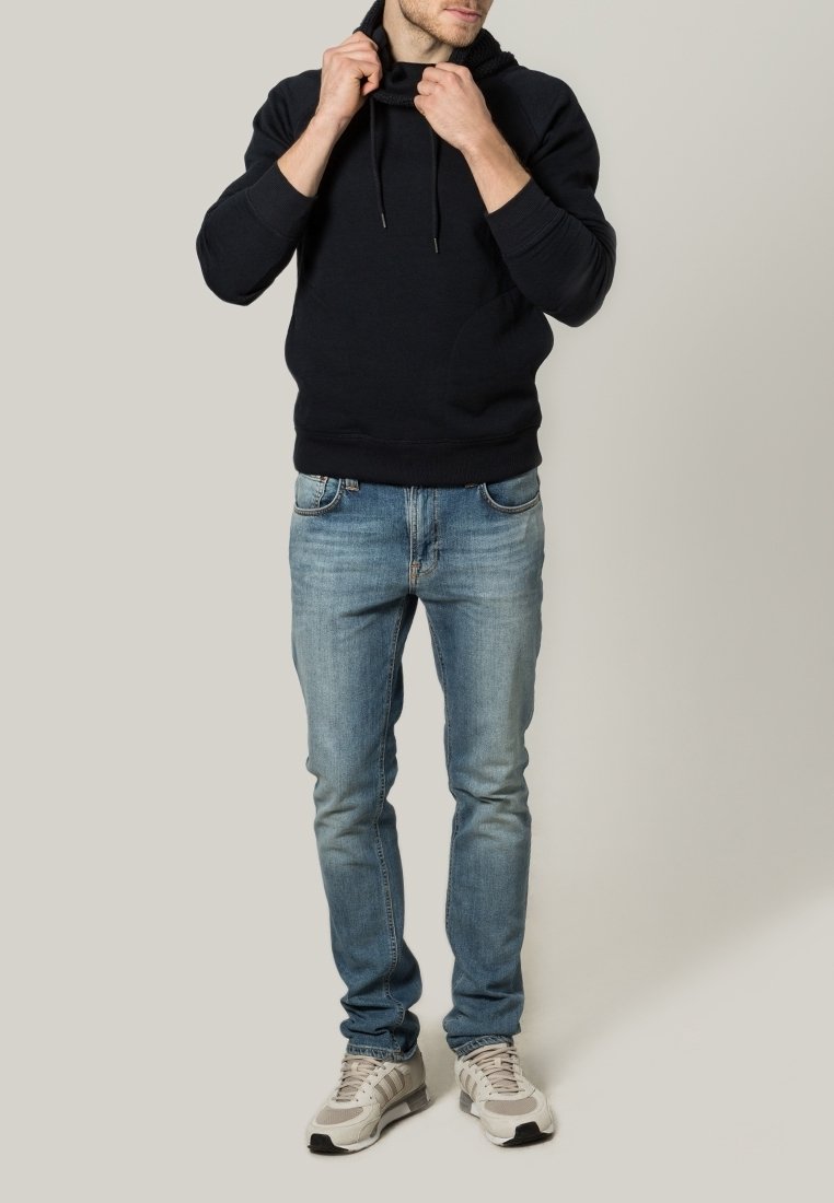 Man adjusting the hood of a black sweatshirt, wearing light blue jeans and white sneakers, standing against a plain light background.