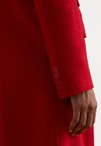 Mâneca unui palton roșu din lână, cu un finisaj texturat și șase butoane asortate de-a lungul manșetei. Design neted și ajustat.