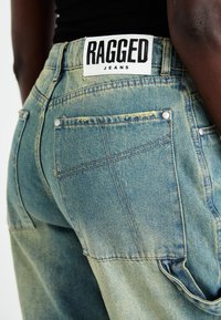 Τζιν παντελόνι σε ξεθωριασμένο ανοιχτό μπλε χρώμα με πίσω τσέπες, χρυσές ραφές και λευκή ετικέτα που γράφει "RAGGED JEANS" κοντά στη μέση.