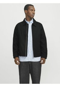 Veste noire zippée avec un col, en tissu lisse. Portée par-dessus une chemise rayée bleu clair, accompagnée d'un pantalon en denim foncé.