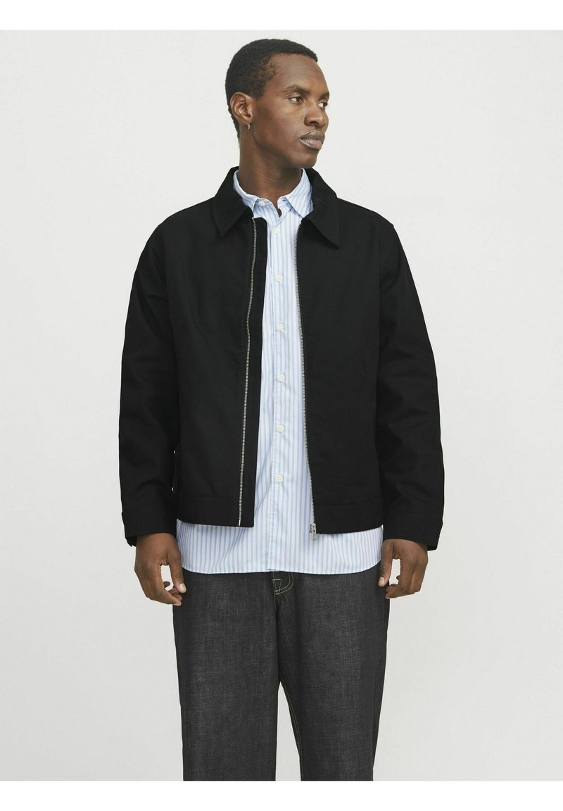 Veste noire zippée avec un col, en tissu lisse. Portée par-dessus une chemise rayée bleu clair, accompagnée d'un pantalon en denim foncé.