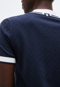 Άνδρας που φοράει κοντομάνικο πουκάμισο σε ναυτικό μπλε με υφή, με λευκή ρίγα στο γιακά και στις μανσέτες, εμφανίζεται από πίσω.