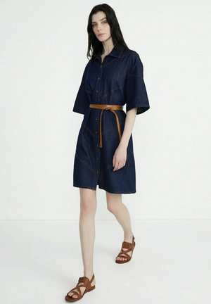 Femme marchant vêtue d'une robe en jean bleu foncé boutonnée avec une ceinture marron et des sandales marron à lanières, sur un fond blanc uni.