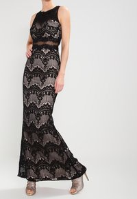 Svart spets maxiklänning med en figurnära livstycke, scalloped fåll och genomskinliga detaljer. Har intrikata blommönster och är ärmlös.