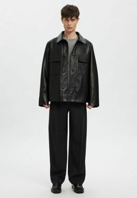 Mustast nahast jakk nööpidega, kahe rinnataskuga, laiemate varrukatega ja klassikalise kraega, kantud halli topi ja tumedate pükste peal.