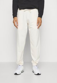 Person som bär vit snörning träningsbyxor och vita sneakers, stående mot en enfärgad vit bakgrund.