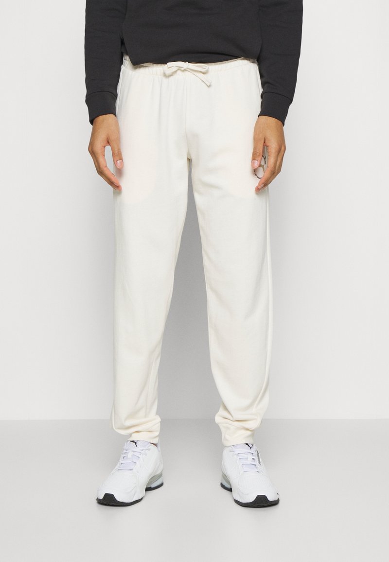 Person som bär vit snörning träningsbyxor och vita sneakers, stående mot en enfärgad vit bakgrund.