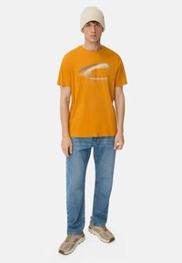 Orange bomuld t-shirt med et stiliseret sølvlogo foran, parret med blå denim jeans og beige sneakers, båret med en cremefarvet strikket beanie.