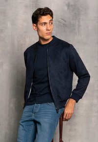 Chaqueta bomber azul marino de tejido suave con puños y cintura acanalados, combinada con una camiseta azul oscura y unos pantalones vaqueros azul claro.