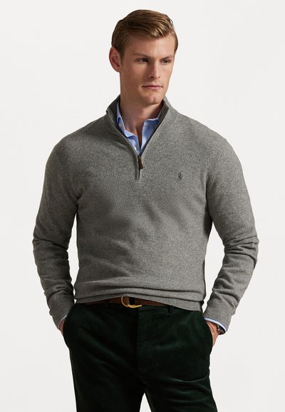 Pull gris à col zippé avec une texture douce, poignets côtelés et un petit logo sur la poitrine, associé à un pantalon en velours côtelé vert foncé.