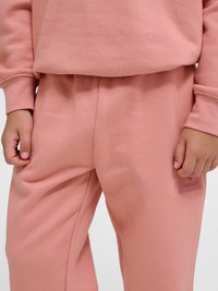 Dziecko ubrane w różowe spodnie dresowe i pasującą bluzę, z widocznymi rękami po bokach oraz prostokątną naszywką z logo na lewej udzie.
