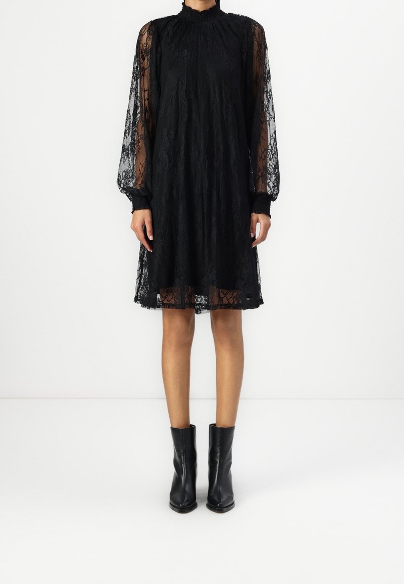 Femme portant une robe noire en dentelle longueur genou avec des manches longues transparentes et des bottines noires, debout devant un fond blanc uni.