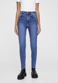 Vaqueros de denim azul ajustados con cintura alta, que cuentan con bolsillos frontales, pasadores para cinturón y cierre de cremallera. Combinados con botas negras de plataforma.